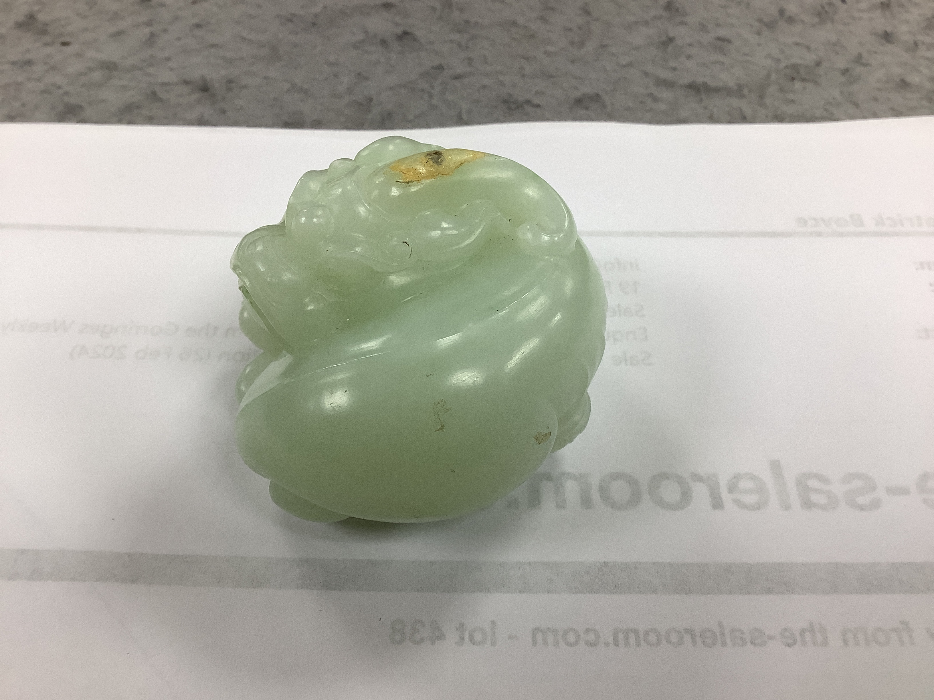 A Chinese celadon jade carving of a lion-dog, in recumbent pose, its crest with a russet inclusion, 5.3cm